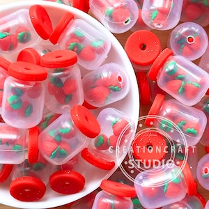 Peut inclure: Une collection de petits bocaux en plastique transparent avec des couvercles rouge vif, contenant chacun des breloques miniatures en forme de fraise rouge avec des feuilles vertes. Les bocaux sont éparpillés dans et autour d'un bol blanc.