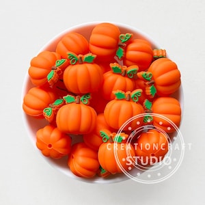 Puede incluir: Una colección de cuentas con forma de calabaza naranja con detalles de hojas verdes, exhibidas en un cuenco blanco. Las cuentas varían en tamaño y forma, algunas con una textura acanalada. La imagen incluye el texto "CREATIONCRAFT STUDIO".