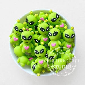 May include: A collection of lime green alien figurines with black eyes, eyelashes, and pink heart-shaped chests. Each alien has small yellow flower accents on its head. The figurines are in a white bowl.