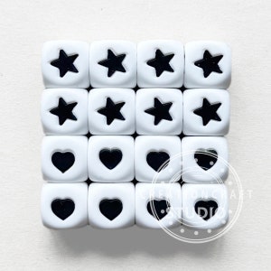 May include: Twenty white silicone beads with black star and heart shapes. The beads are arranged in a grid pattern.