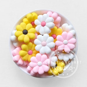 Pode incluir: Uma tigela de pingentes de silicone em forma de flor em rosa, branco e amarelo. Os pingentes são perfeitos para adicionar um toque de fantasia a joias, chaveiros ou outros artesanatos.