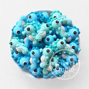 Peut inclure: Un ensemble d'ornements en forme d'hippocampes bleus et turquoise, avec des détails blancs et des yeux noirs. Les hippocampes sont dans un bol blanc. Le texte "CREATIONCRAFT STUDIO" est visible.