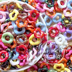 May include: A bowl filled with colourful iridescent ceramic beads in various shapes, including rings, ovals, and swirls. The beads have a glossy finish and are perfect for jewellery making or crafting.