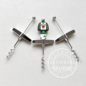 May include: Three silver wine bottle openers with a Christmas sweater design. The sweater is red, green, and white with candy canes and holly.