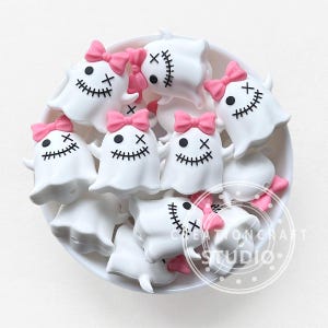 Pode incluir: Uma coleção de amuletos brancos em forma de fantasma com sorrisos costurados, olhos cruzados pretos e laços rosa. Os amuletos estão dispostos numa tigela branca, perfeitos para o Halloween ou projetos de artesanato.