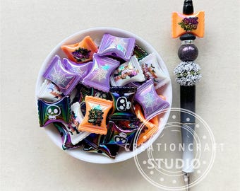 Może przedstawiać: Miska wypełniona kolorowymi, halloweenowymi charmsami w kształcie cukierków. Charms mają kształt opakowań cukierków i przedstawiają wzory takie jak czaszki, pajęczyny i napis "Trick or Treat".