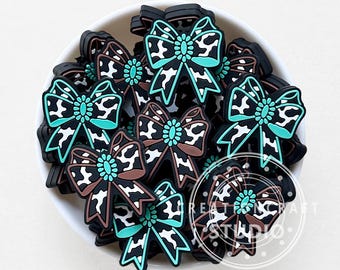 May include: A collection of black and white cow print bow-shaped embellishments with teal accents, displayed in a white bowl. The bows have a central jewel-like detail. These decorative bows are suitable for various craft projects.