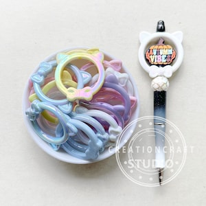 May include: A white bowl filled with iridescent cat-shaped rings in various pastel colors. A black and white pen with a white pumpkin charm that says "Autumn Vibes" is next to the bowl.