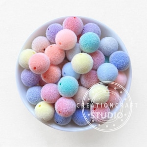 Op de afbeelding: Een witte kom gevuld met kleurrijke, fuzzy pom-pom kralen in tinten roze, paars, blauw en geel. De kralen zijn rond met een klein gaatje. De afbeelding bevat de tekst "CREATIONCRAFT STUDIO".