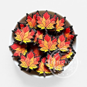 May include: A collection of small, flat maple leaf-shaped charms in a white bowl. Each charm features a gradient of red, orange, and yellow segments outlined in black. The text CREATIONCRAFT STUDIO is visible in a circular watermark on the lower right.
