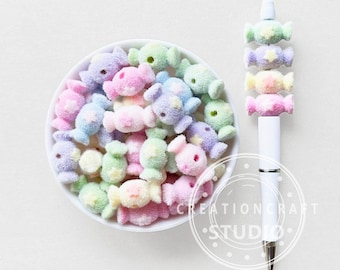 May include: A white ceramic bowl filled with pastel-colored candy-shaped decorations. A white pen is adorned with similar candy-shaped embellishments. The candies are in shades of pink, blue, green, and yellow, with a star detail.