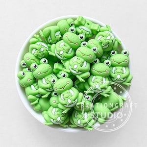 Peut inclure: Un bol blanc rempli d'ornements en forme de grenouilles vert clair. Chaque grenouille a un visage souriant, et certaines tiennent une plus petite grenouille. L'image comprend le texte "CREATIONCRAFT STUDIO".