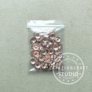 May include: A clear plastic bag filled with rose gold-colored metal beads. The beads have a faceted, textured surface and a circular shape with a central hole. The bag is sealed and sits on a light gray surface.