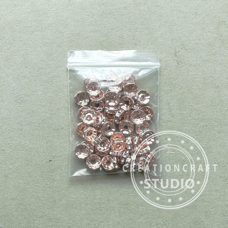 May include: A clear plastic bag filled with rose gold-colored metal beads. The beads have a faceted, textured surface and a circular shape with a central hole. The bag is sealed and sits on a light gray surface.