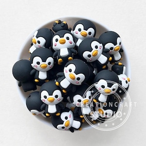 Pode incluir: Uma tigela branca cheia de inúmeras contas em forma de pinguim pretas, brancas e amarelas. Cada conta apresenta um desenho de pinguim de desenho animado com corpo preto, barriga branca e bico amarelo. O texto "CREATIONCRAFT STUDIO" é visível.