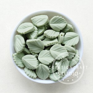 May include: A white bowl filled with pale green leaf-shaped beads. The beads have detailed veining and a hole for stringing. The image includes the text "CREATIONCRAFT STUDIO" in a circular design. These beads are suitable for jewellery making and craft projects.