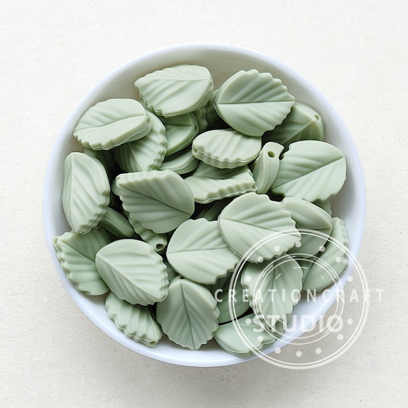 May include: A white bowl filled with pale green leaf-shaped beads. The beads have detailed veining and a hole for stringing. The image includes the text "CREATIONCRAFT STUDIO" in a circular design. These beads are suitable for jewellery making and craft projects.