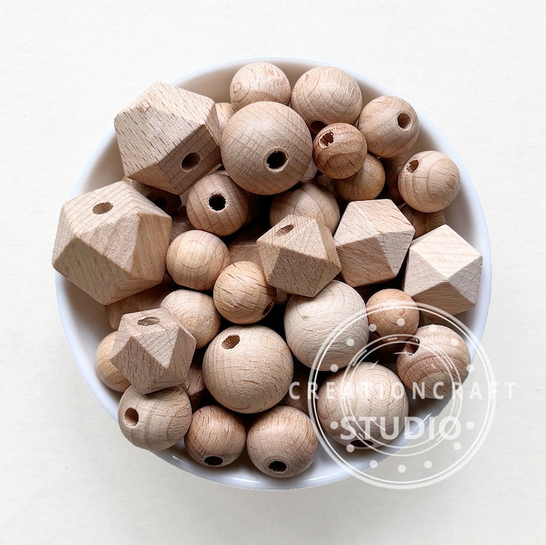 May include: A bowl filled with natural wood beads in various shapes and sizes. The beads are a light brown color and have a smooth, polished finish. Some of the beads are round, while others are geometric shapes, such as squares, triangles, and hexagons. The beads are perfect for crafting jewelry, garlands, or other decorative projects.