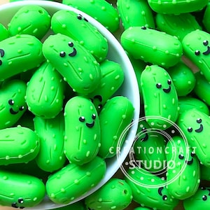 May include: A close-up of numerous bright green, cartoon-style pickle charms, each with a smiling face and small bumps. Some are in a white bowl, others scattered around. The text "CREATIONCRAFT STUDIO" is visible.
