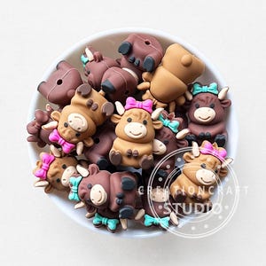 Puede incluir: Un conjunto de adornos con forma de vaca en marrón y beige, con lazos rosas y azules, dispuestos en un cuenco blanco. Las figuras de vaca tienen caras sonrientes y están hechas de un material liso. La imagen está bien iluminada.