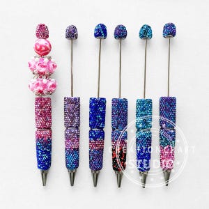 May include: An assortment of decorative pens with sparkling, jewel-toned embellishments. The pens feature a variety of colors, including pink, purple, blue, and teal, with silver-colored metal tips. Some pens have beaded accents, while others have a solid, glittery design.