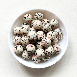 May include: A bowl of white silicone beads with a black, pink, and blue terrazzo pattern. The beads are approximately 2 cm in diameter.
