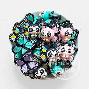 Pode incluir: Uma coleção de pequenos pingentes de borboleta planos em estilo cartoon dentro de uma tigela branca. Cada pingente apresenta um personagem com olhos grandes e asas coloridas em tons de turquesa, rosa, roxo, amarelo e laranja, contornados.