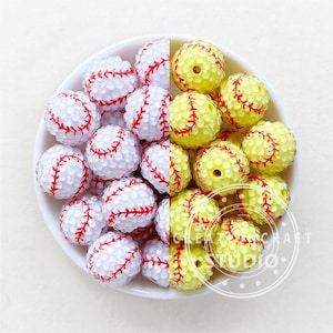 May include: A bowl filled with white and yellow baseball-themed beads. The beads are decorated with red stitching and have a textured surface.