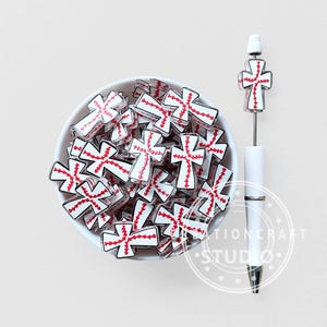 May include: A white bowl filled with multiple cross-shaped beads featuring a white surface and red stitched detailing resembling baseball seams. Beside the bowl is a white pen topped with a matching cross-shaped bead with red stitching.