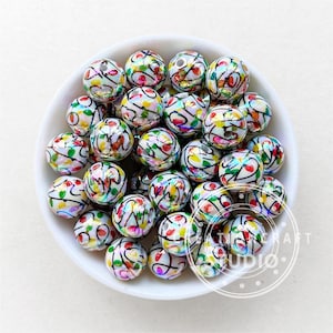 Puede incluir: Un conjunto de cuentas redondas blancas con diseños abstractos coloridos en un cuenco blanco. Las cuentas presentan patrones rojos, verdes, amarillos y negros. Las cuentas son probablemente para la fabricación de joyas o manualidades.