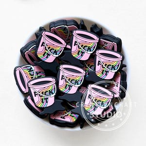 Pode incluir: Uma coleção de pingentes rosa em forma de balde com um design de arco-íris e a palavra "F*CK IT" em branco. Os pingentes estão em uma base preta e em uma tigela branca.