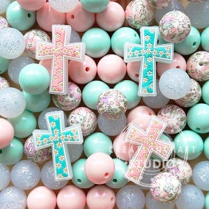 May include: A collection of colorful beads with floral patterns and cross charms. The beads are in shades of pink, mint green, and white. Some beads have glitter.
