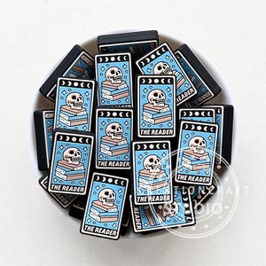 Może przedstawiać: Stos niebieskich i czarnych prostokątnych zawieszek z motywem czaszki i stosu książek. Zawieszki mają napis "The Reader".