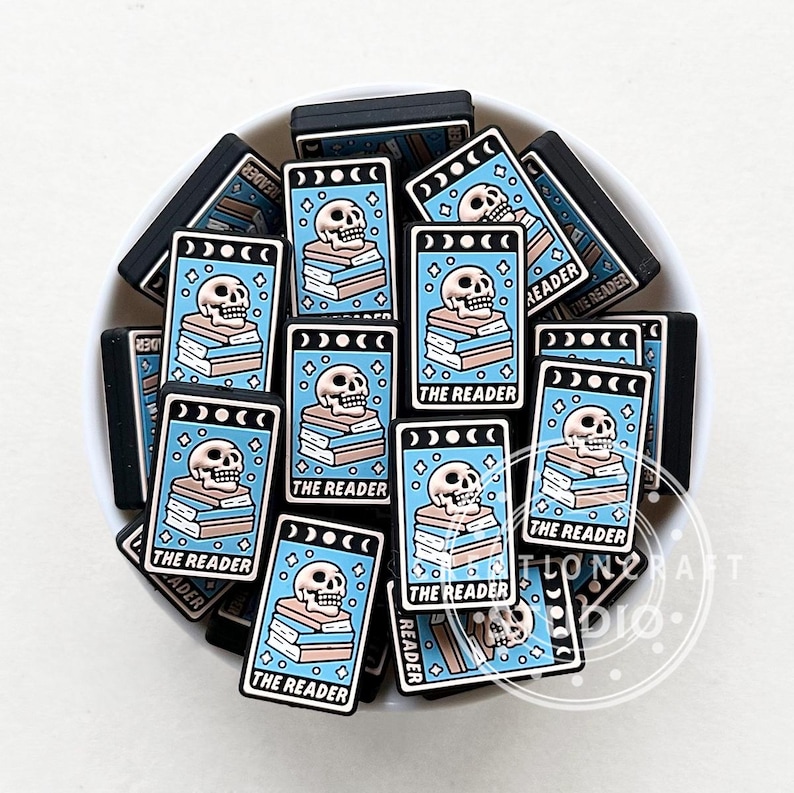 Może przedstawiać: Stos niebieskich i czarnych prostokątnych zawieszek z motywem czaszki i stosu książek. Zawieszki mają napis "The Reader".