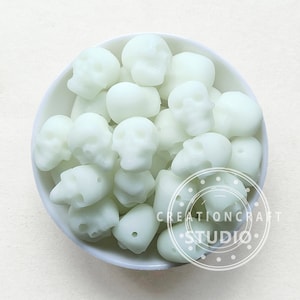May include: A white bowl filled with glow-in-the-dark skull-shaped beads. The beads are a pale green color and have a matte finish.