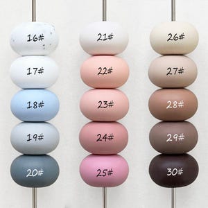 Puede incluir: Un conjunto de 15 cuentas redondas de cerámica en varios tonos de blanco, rosa, marrón y gris. Las cuentas están dispuestas en tres columnas de cinco cuentas cada una, con cada cuenta etiquetada con un número del 16 al 30.