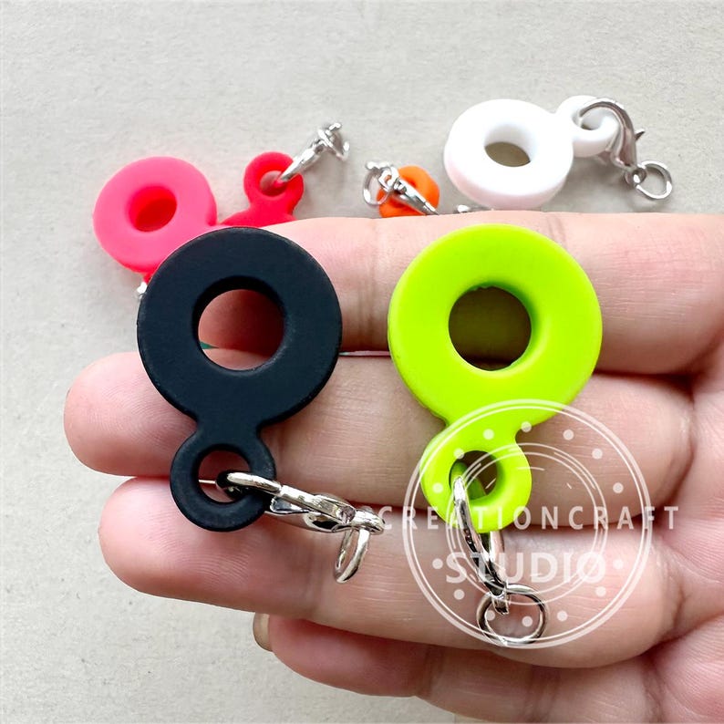May include: Assortment of colorful silicone keychains with silver metal rings and clasps. The keychains are shaped like a circle with a smaller circle attached to the side. The colors include black, green, pink, and white.