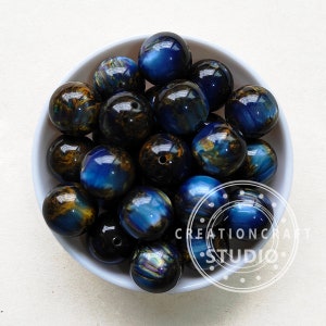 Op de afbeelding: Een kom met blauwe en gouden tijgeroog kralen. De kralen zijn rond en hebben een glanzende, iriserende afwerking.