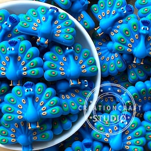 以下が含まれることがあります： 緑とオレンジのアクセントが付いた、鮮やかな青色の孔雀型の装飾品のコレクション。装飾品は白いボウルに入っており、周囲に散らばっています。「CREATIONCRAFT STUDIO」の文字が見えます。
