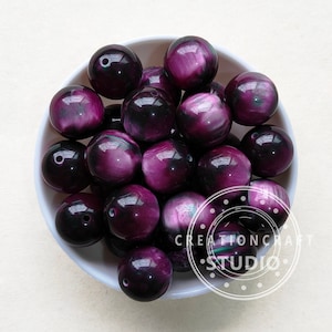 Op de afbeelding: Een kom gevuld met ronde, iriserende kralen in paars en zwart. De kralen hebben een glanzende afwerking en zijn perfect voor het maken van sieraden.