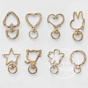 May include: A selection of gold-coloured metal swivel clasps in diverse shapes, such as a heart, star, bunny, and flower. Each clasp features a D-ring and a swivel hook, ideal for keychains or accessories.