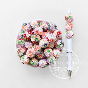 Könnte beinhalten: Eine weiße Schüssel gefüllt mit bunten Blumenperlen. Ein weißer Stift ist mit ähnlichen Blumenperlen verziert. Die Perlen sind in den Farbtönen Rot, Rosa, Gelb, Grün und Lila gehalten. Der Stift hat eine silberne Spitze.