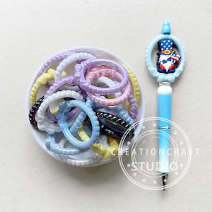 May include: A white bowl filled with a variety of colorful, iridescent plastic rings. A blue pen with a red, white, and blue gnome design on the top is next to the bowl. The pen has the text "CREATIONCRAFT STUDIO" on the side.