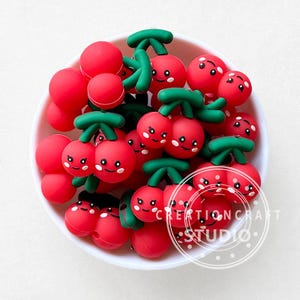 May include: A white bowl filled with red cherry-shaped beads with green stems and smiling faces. The beads are a mix of single cherries and pairs. The text "CREATION CRAFT STUDIO" is visible on the bottom right.