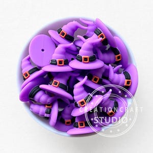 May include: A collection of purple witch hat charms with black and orange accents, displayed in a white bowl. The charms have a classic witch hat shape, perfect for Halloween-themed crafts. The image includes the text "CREATION CRAFT STUDIO".
