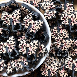 Puede incluir: Una colección de dijes de lazo con estampado de leopardo negro y beige con detalles de estetoscopio rosa. Los dijes están en un cuenco blanco y esparcidos sobre una superficie de madera. Los dijes son probablemente para zapatos o accesorios.