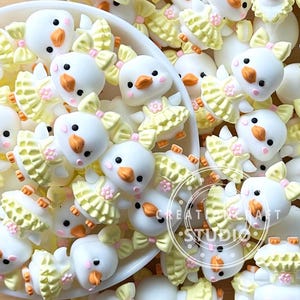 May include: A collection of white chick-shaped embellishments with orange beaks and black eyes. Some chicks wear yellow dresses with pink flower accents. The chicks have yellow bows and are scattered in a white bowl. The image has the text "CREATE CRAFT STUDIO".