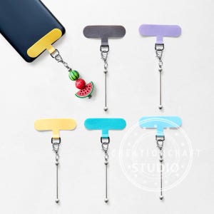 Puede incluir: Una colección de accesorios de agarre para teléfonos de colores. Los agarres son de varios colores, incluyendo gris, morado, amarillo, turquesa y azul claro. Cada agarre tiene una cadena plateada con una pequeña bola al final. Un agarre tiene un dije con sandía y fruta.