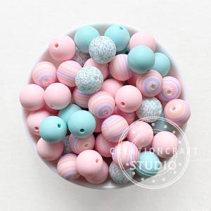 Peut inclure: Un assortiment de perles en silicone aux couleurs pastel dans un bol blanc. Les perles sont roses, bleues et blanches, certaines avec des motifs rayés ou tachetés. Les perles sont rondes et percées.