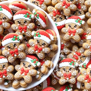 Könnte beinhalten: Eine Sammlung von Lebkuchenmann-förmigen Ornamenten. Jedes Ornament ist hellbraun mit weißen Zuckergussdetails, einer roten Fliege und einem rot-weißen Weihnachtsmannhut. Die Ornamente befinden sich in einer weißen Schüssel und sind auf einer Holzoberfläche verstreut.